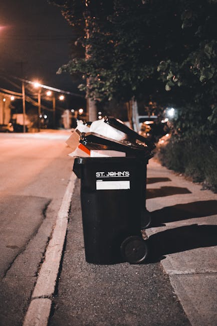 A person places a crumpled brown paper bag into a white waste bin labeled 'RUBBISH' situated indoors, likely in a residential setting such as a flat or estate. The person's arm extends from the left s